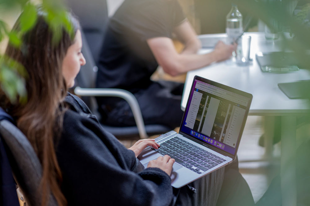 Junge Frau im Hoodie tippt auf MacBook Air, verschwommene Pflanzen im Vordergrund, sitzender Kollege im Hintergrund