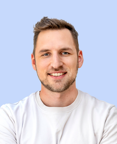 Junger Mann mit kurzem braunem Haar und leichtem Bartstoppel trägt helles Sweatshirt, lächelnd vor pastellblauem Hintergrund