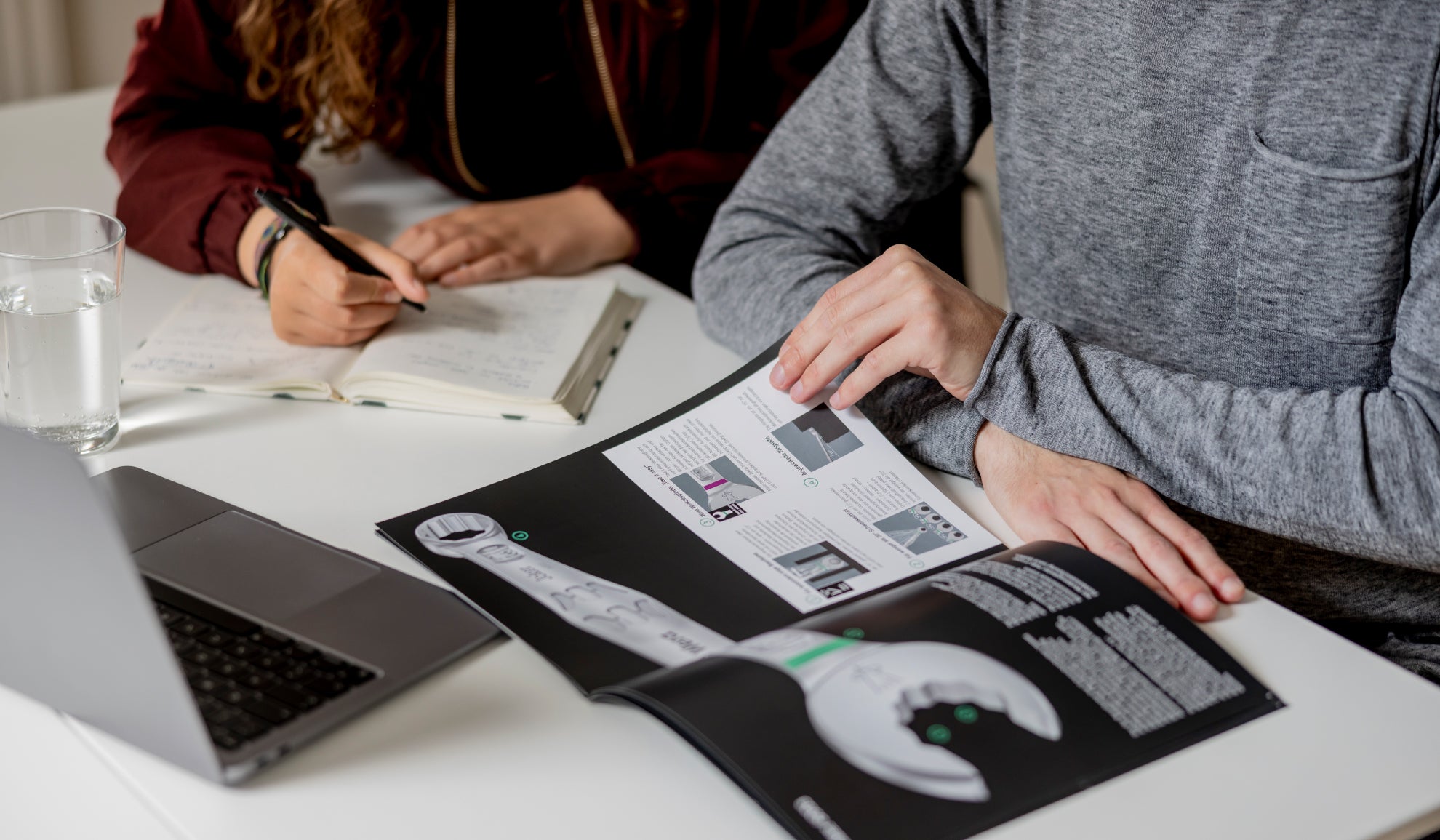 zwei Personen am Tisch schreiben Notizen in aufgeklapptem Notizbuch und blättern in geöffnetem Prospekt neben Laptop und Glas Wasser