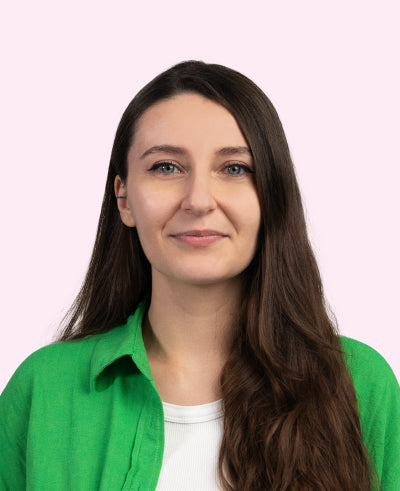 Frau mit langen braunen Haaren und grüner Bluse vor pastellrosa Hintergrund lächelt dezent