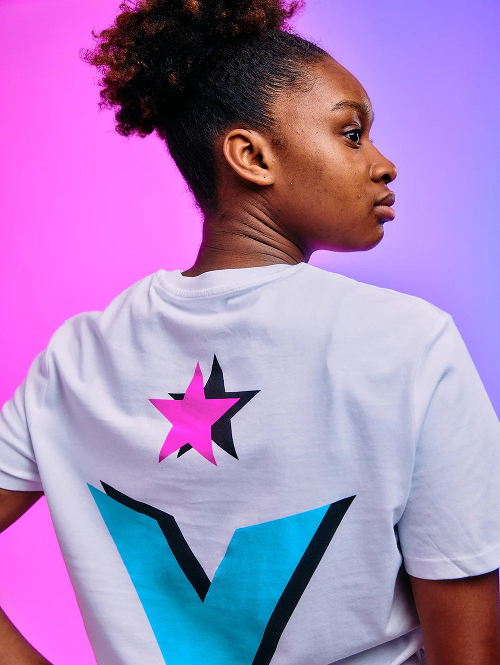 Junge schwarze Frau im Profil mit weißem T-Shirt und buntem Stern-V-Logo vor rosa-violettem Hintergrund