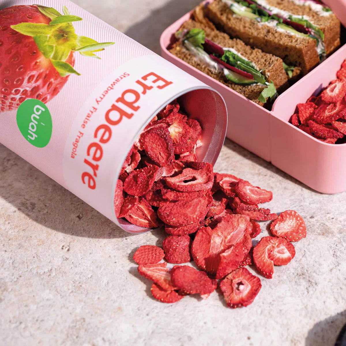 rosa Zylinder mit roten gefriergetrockneten Erdbeerscheiben neben rosa Lunchbox mit Vollkornbrotsandwiches und Blattgemüse