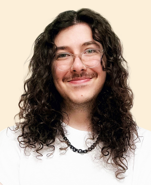 A smiling person with long curly brown hair, wearing clear glasses, a white shirt, and a black chain necklace, standing against a light beige background.