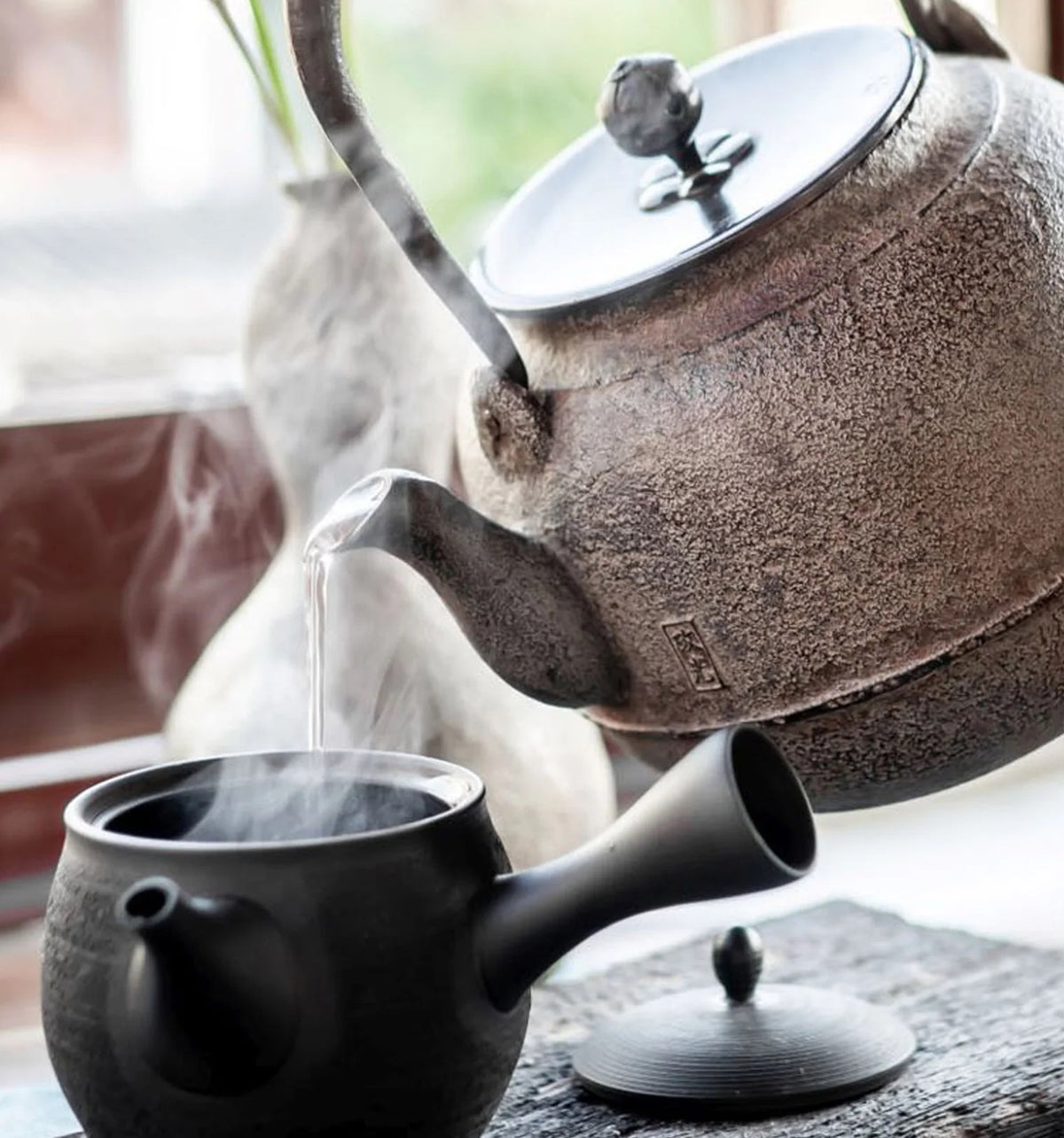 Gusseiserne Teekanne gießt heißes Wasser in abgesetzte schwarze Kyusu-Teekanne auf Holztablett