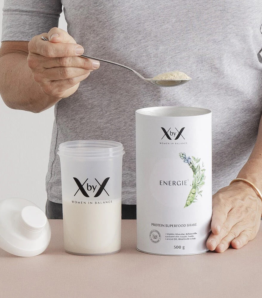 Hände halten Löffel mit weißem Proteinpulver über geöffnetem Shake-Becher mit Milch neben XbyX-Energieshake-Dose