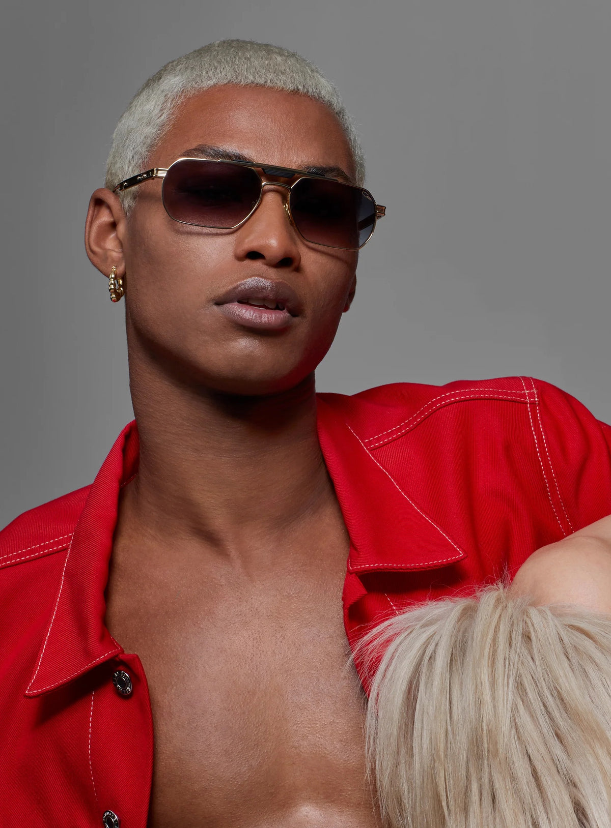 androgynes Modell mit platinblondem Kurzhaarschnitt, goldgerahmten Sonnenbrillen und offener roter Jacke
