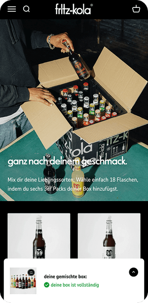 Person greift in geöffnete Kartonbox mit bunten fritz-kola Flaschen vor dunklem Hintergrund